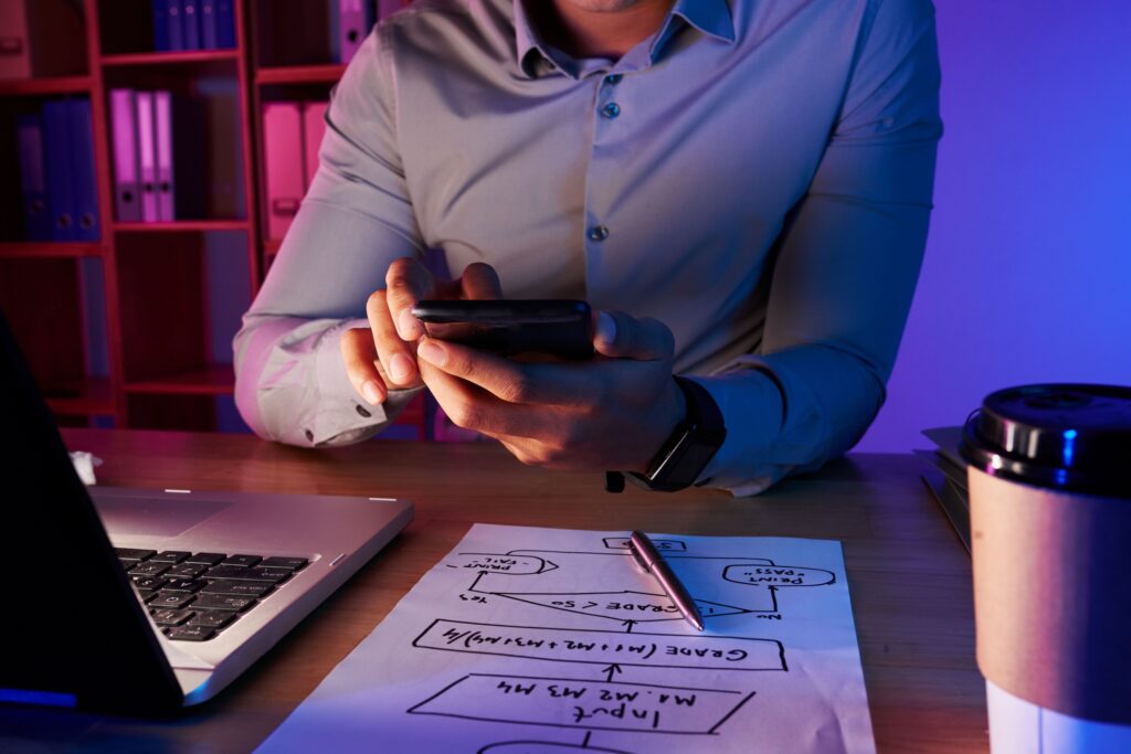 Homme d'affaires en chemise consultant son smartphone devant un plan de marketing digital et un ordinateur portable.
